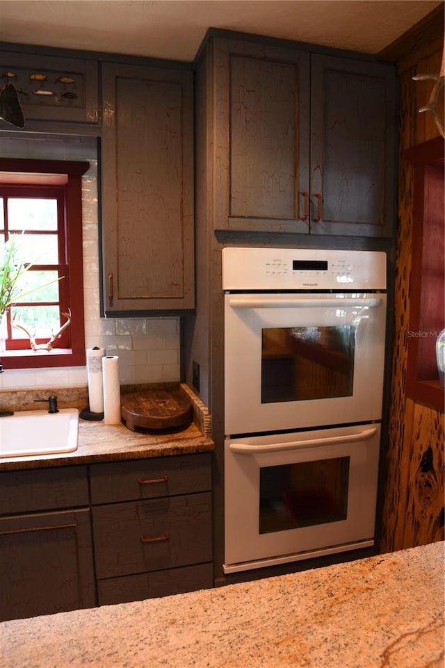 kitchen featuring light stone counters, backsplash, stainless steel double oven, dark brown cabinets, and sink