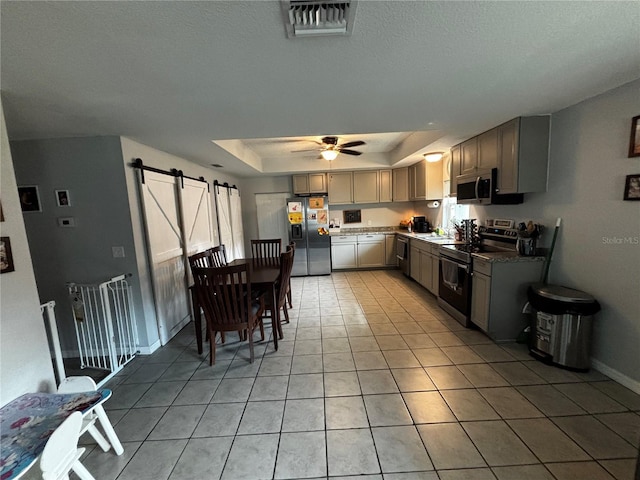 kitchen with gray cabinetry, stainless steel appliances, a barn door, a tray ceiling, and light tile patterned floors