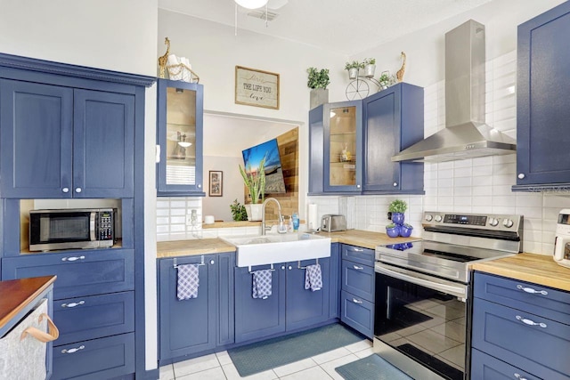 kitchen with butcher block counters, glass insert cabinets, stainless steel appliances, wall chimney range hood, and a sink