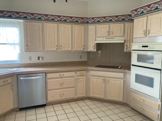 kitchen with white double oven, black electric cooktop, light tile floors, stainless steel dishwasher, and tile counters