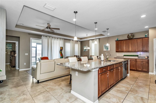 kitchen featuring a tray ceiling, ceiling fan, sink, an island with sink, and light stone counters