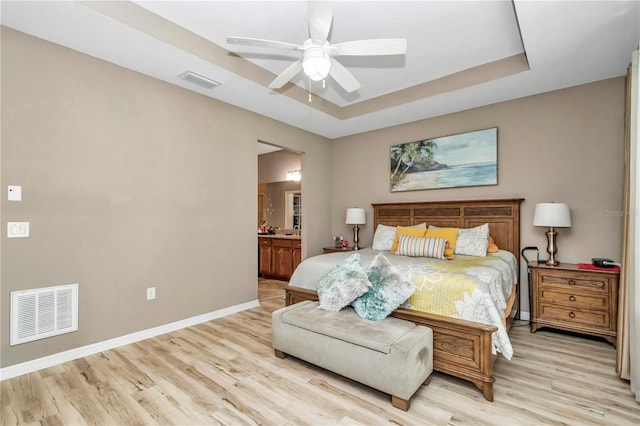 bedroom with a tray ceiling, ensuite bathroom, ceiling fan, and light wood-type flooring