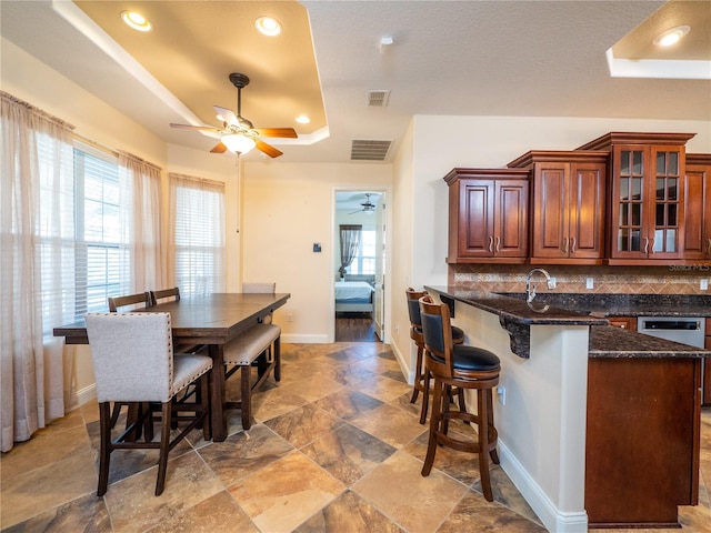 kitchen with backsplash, a tray ceiling, ceiling fan, dark stone countertops, and a breakfast bar