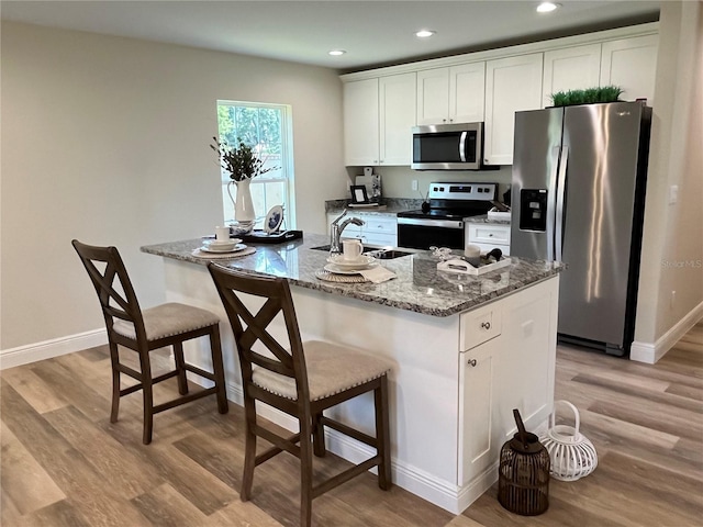 kitchen with dark stone countertops, a center island with sink, stainless steel appliances, light wood-type flooring, and white cabinets