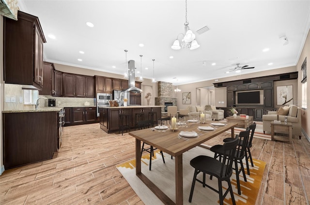 dining space with ceiling fan with notable chandelier, a stone fireplace, ornamental molding, and light hardwood / wood-style flooring