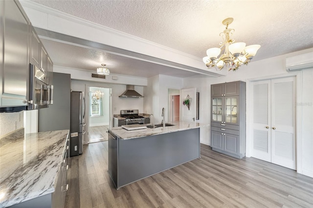 kitchen with appliances with stainless steel finishes, light stone counters, wall chimney exhaust hood, sink, and an island with sink