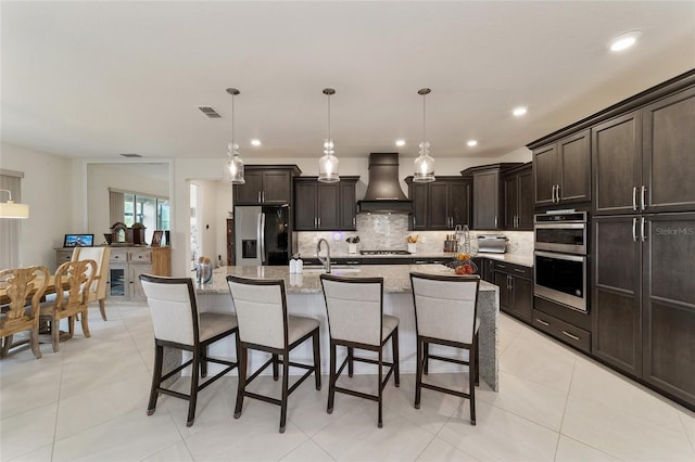 kitchen with appliances with stainless steel finishes, a breakfast bar, custom range hood, a center island with sink, and hanging light fixtures