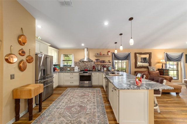 kitchen featuring appliances with stainless steel finishes, a breakfast bar, wall chimney range hood, decorative light fixtures, and tasteful backsplash