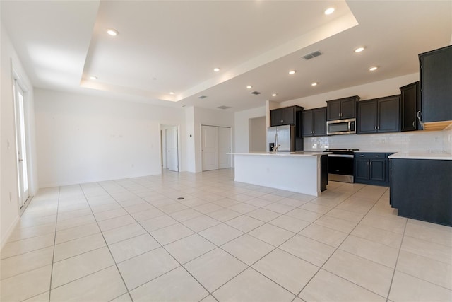 kitchen with light tile patterned flooring, backsplash, a tray ceiling, stainless steel appliances, and a kitchen island with sink