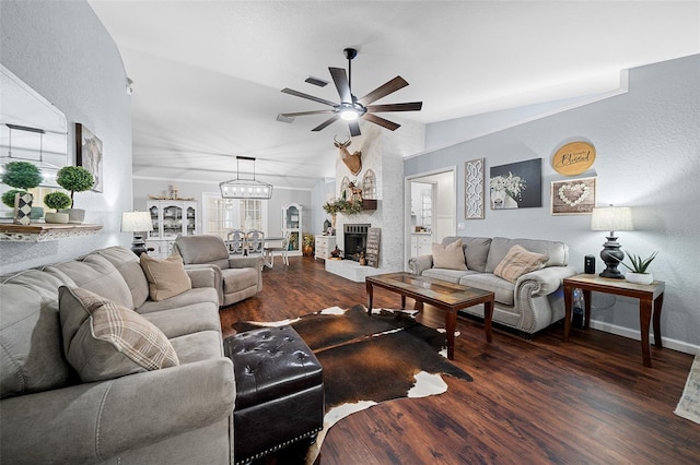 living room with a brick fireplace, ceiling fan with notable chandelier, vaulted ceiling, and dark wood-type flooring