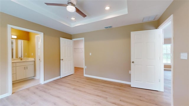 unfurnished bedroom with sink, ensuite bath, a walk in closet, light wood-type flooring, and a tray ceiling