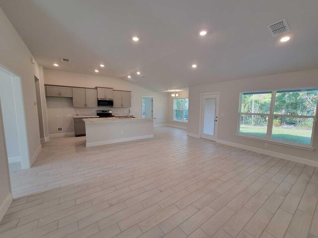 kitchen featuring appliances with stainless steel finishes, light stone counters, gray cabinetry, vaulted ceiling, and a center island with sink