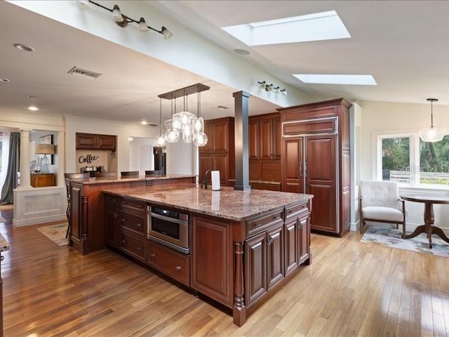 kitchen with vaulted ceiling with skylight, decorative light fixtures, light wood-type flooring, and a kitchen island with sink