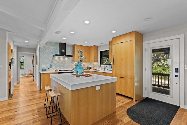 kitchen featuring wall chimney range hood, sink, light hardwood / wood-style flooring, beam ceiling, and a kitchen island