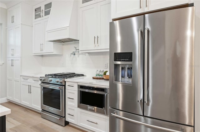 kitchen with appliances with stainless steel finishes, tasteful backsplash, custom range hood, white cabinets, and light hardwood / wood-style floors