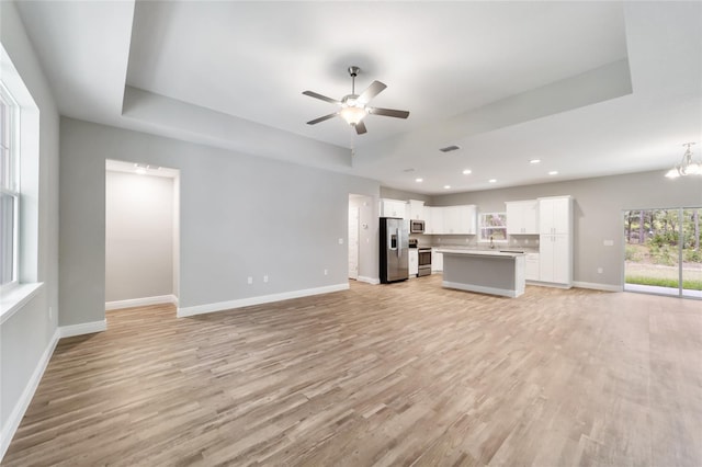 unfurnished living room with a raised ceiling, ceiling fan with notable chandelier, and light wood-type flooring