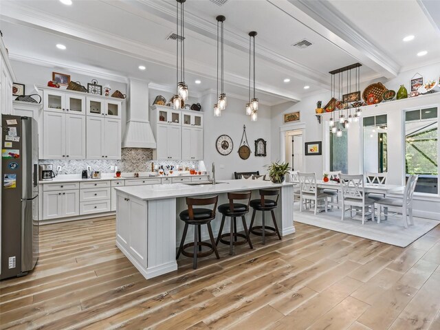 kitchen featuring white cabinets, beamed ceiling, a center island with sink, custom range hood, and stainless steel refrigerator