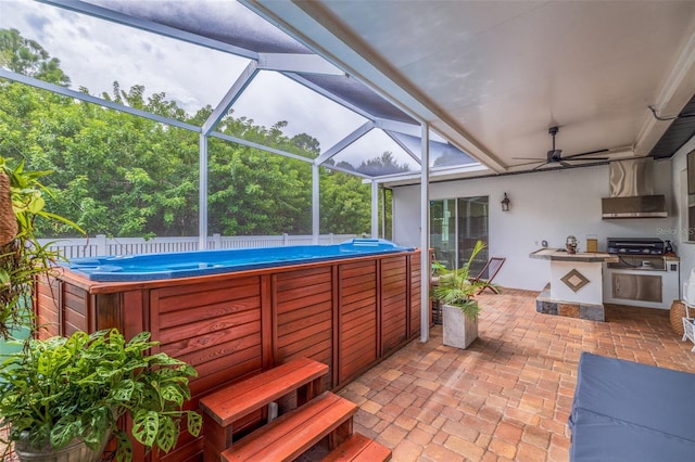 view of patio with a hot tub, fence, glass enclosure, an outdoor kitchen, and a ceiling fan