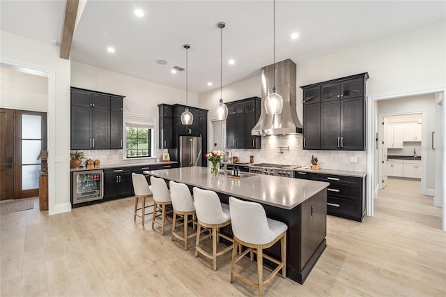 kitchen featuring stainless steel appliances, wall chimney range hood, beverage cooler, an island with sink, and pendant lighting