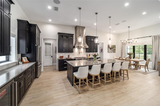 kitchen with a large island with sink, wine cooler, wall chimney exhaust hood, decorative light fixtures, and a kitchen bar