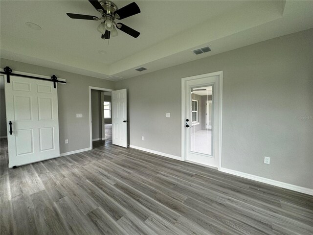 unfurnished bedroom featuring wood-type flooring, a tray ceiling, a barn door, and ceiling fan