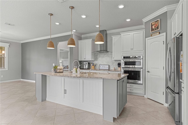 kitchen with wall chimney exhaust hood, appliances with stainless steel finishes, a kitchen island with sink, hanging light fixtures, and light stone counters