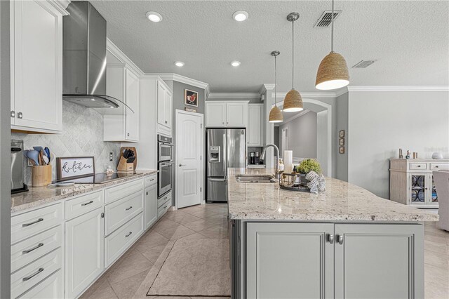 kitchen with pendant lighting, stainless steel appliances, an island with sink, white cabinets, and wall chimney range hood