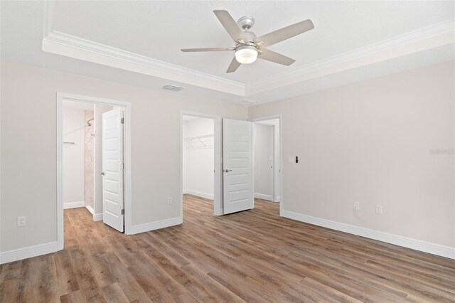 unfurnished bedroom featuring a walk in closet, wood-type flooring, ornamental molding, ceiling fan, and a tray ceiling