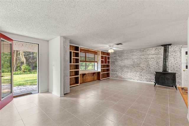 unfurnished living room featuring a textured ceiling, ceiling fan, brick wall, light tile patterned floors, and a wood stove