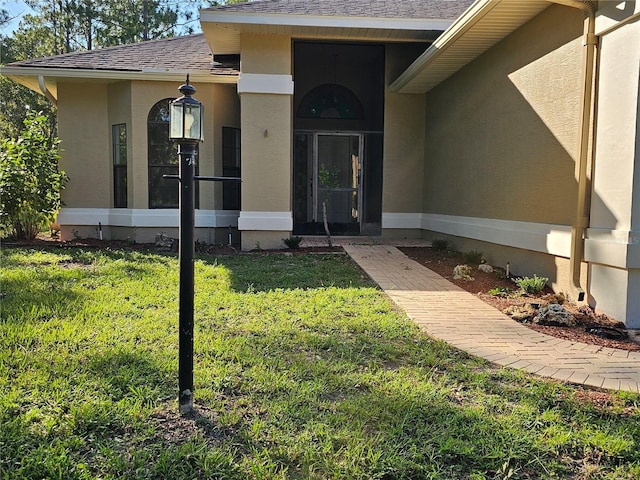 view of exterior entry featuring roof with shingles, a lawn, and stucco siding