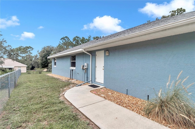 view of exterior entry featuring stucco siding, a shingled roof, fence, and a yard