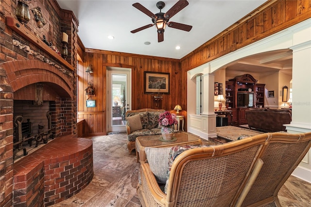 tiled living room with ceiling fan, wood walls, a brick fireplace, decorative columns, and crown molding