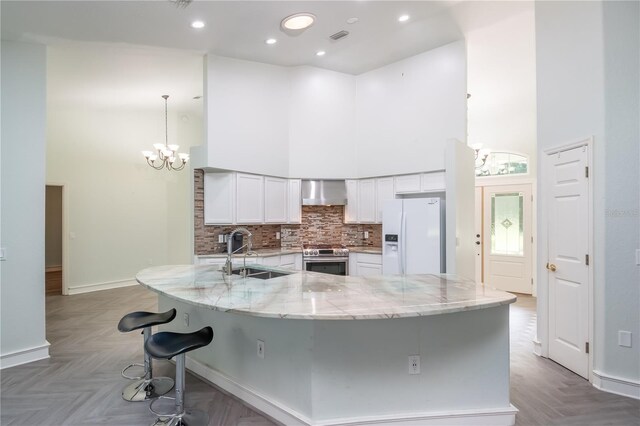 kitchen featuring white refrigerator with ice dispenser, stainless steel range with electric cooktop, sink, a towering ceiling, and wall chimney exhaust hood