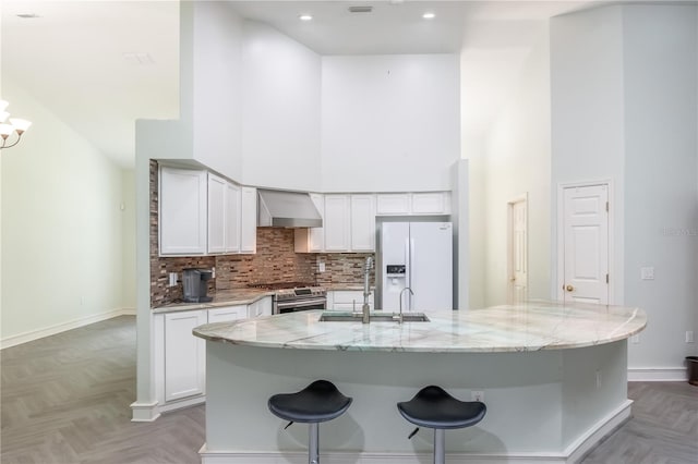 kitchen featuring white refrigerator with ice dispenser, light stone countertops, wall chimney exhaust hood, stainless steel range oven, and a high ceiling