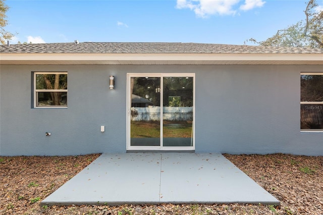 entrance to property featuring a shingled roof, a patio, and stucco siding
