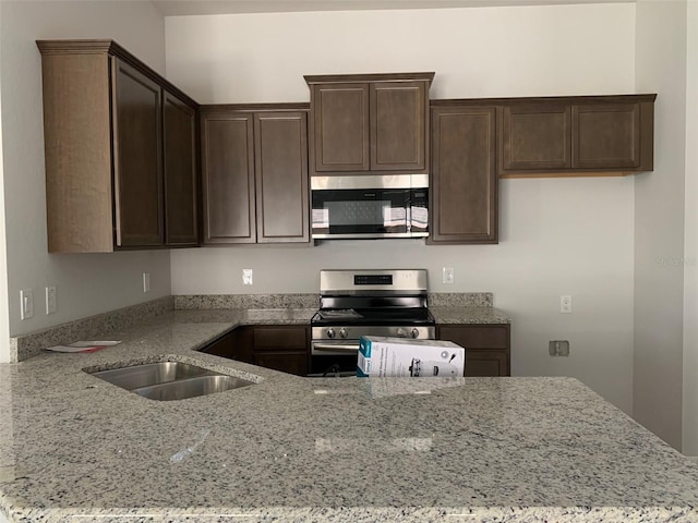 kitchen featuring dark brown cabinetry, stainless steel appliances, a peninsula, a sink, and light stone countertops