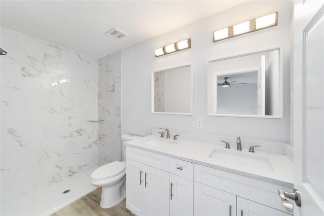 bathroom featuring wood-type flooring, a tile shower, double vanity, ceiling fan, and toilet