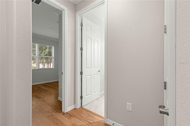 hallway featuring ornamental molding, light wood-type flooring, a textured wall, and baseboards