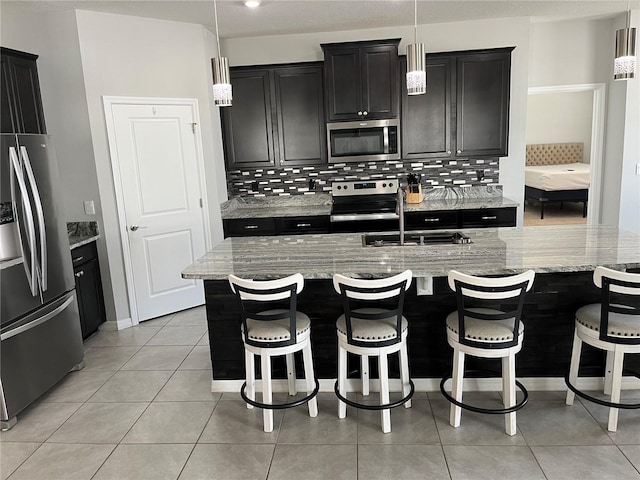 kitchen featuring a center island with sink, appliances with stainless steel finishes, tasteful backsplash, hanging light fixtures, and light stone counters