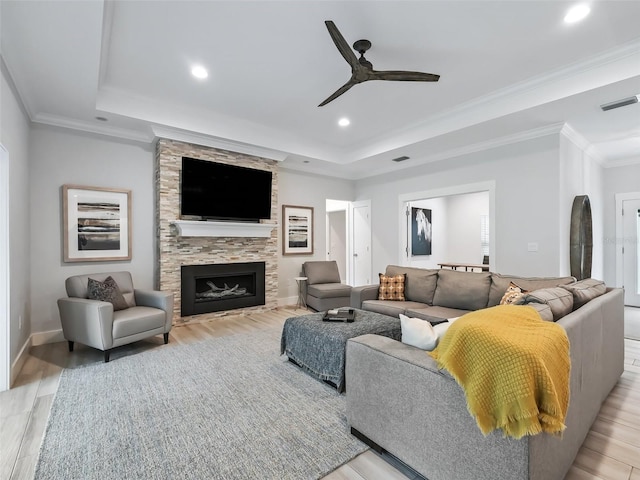 living room with crown molding, light hardwood / wood-style flooring, a raised ceiling, a stone fireplace, and ceiling fan