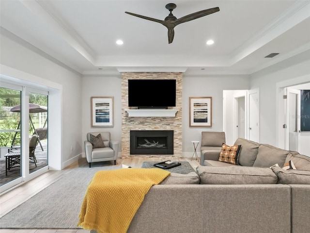 living room with a tray ceiling, ceiling fan, ornamental molding, and a stone fireplace