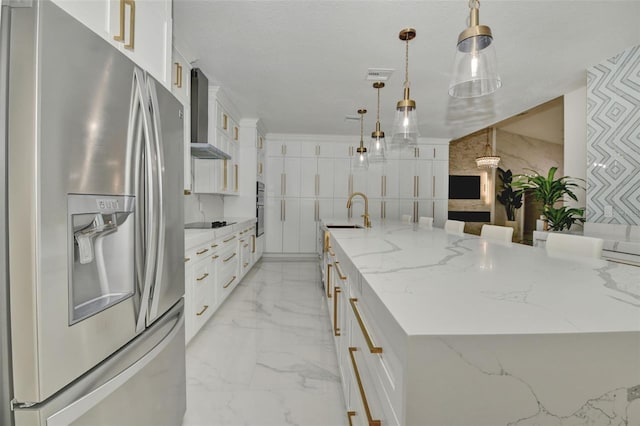 kitchen featuring a large island with sink, white cabinets, stainless steel appliances, decorative light fixtures, and wall chimney range hood