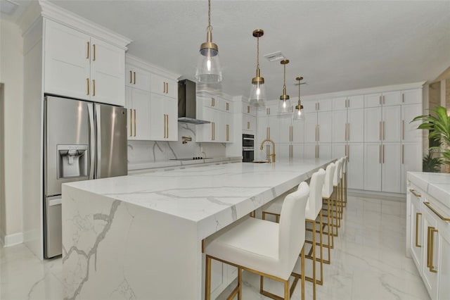 kitchen with pendant lighting, white cabinets, a large island with sink, wall chimney range hood, and appliances with stainless steel finishes