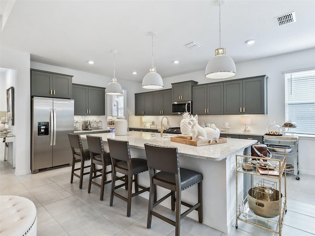 kitchen with decorative light fixtures, appliances with stainless steel finishes, light stone counters, and a kitchen island with sink