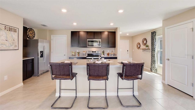 kitchen with a kitchen island with sink, stainless steel appliances, a kitchen breakfast bar, light stone countertops, and dark brown cabinetry