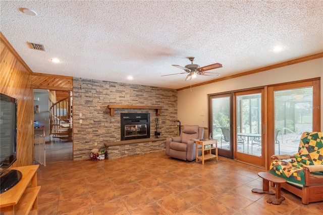 living room featuring a textured ceiling, wood walls, a stone fireplace, ornamental molding, and ceiling fan