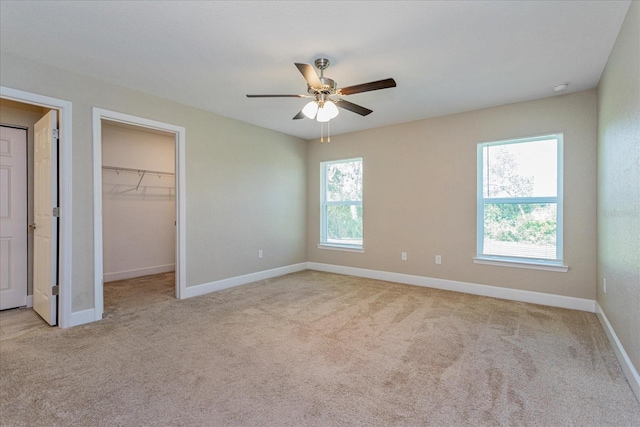 unfurnished bedroom with multiple windows, a walk in closet, light colored carpet, and ceiling fan