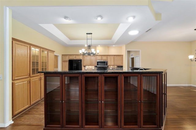 kitchen with black fridge, a notable chandelier, a center island with sink, a raised ceiling, and dark hardwood / wood-style flooring