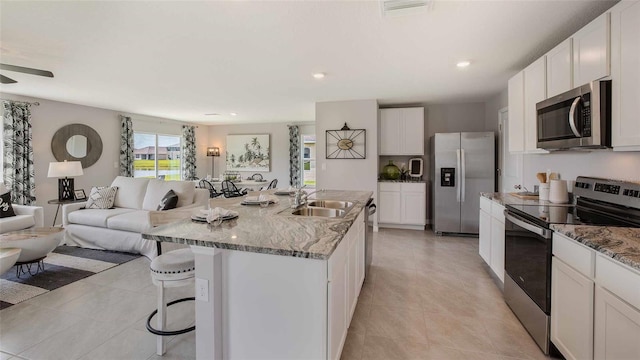 kitchen featuring appliances with stainless steel finishes, light stone countertops, ceiling fan, a center island with sink, and sink