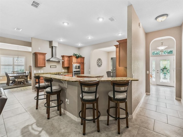 kitchen featuring wall chimney range hood, appliances with stainless steel finishes, light stone countertops, a kitchen bar, and kitchen peninsula